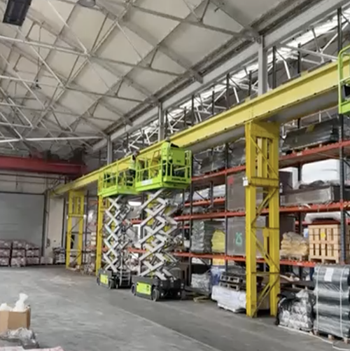 Warehouse interior with yellow industrial hoist and green scissor lifts near stocked shelves.